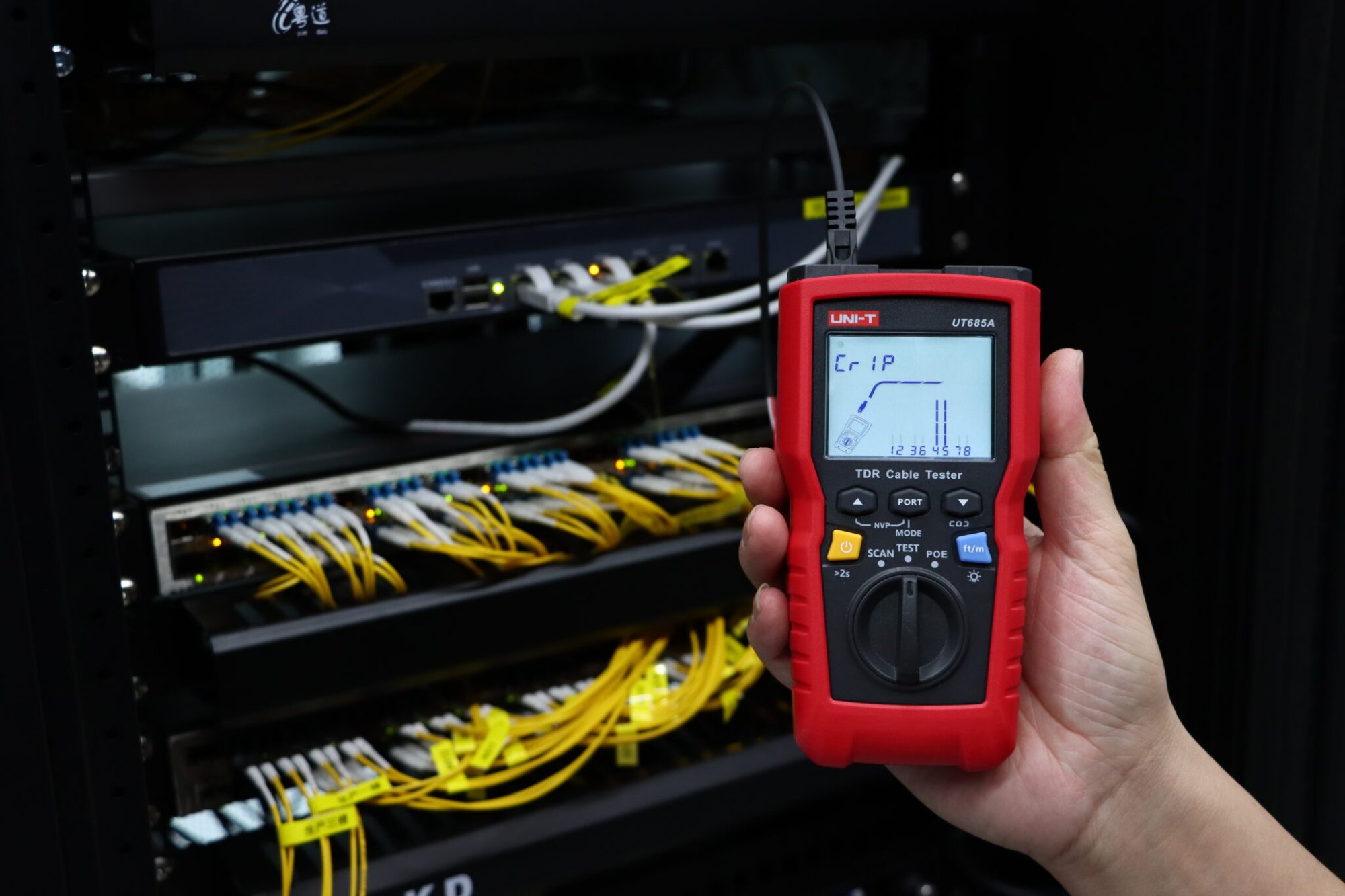 UNI-T UT685A Kabeltester in Servicetechnikerhand vor Switch und Glasfaserverkabelung